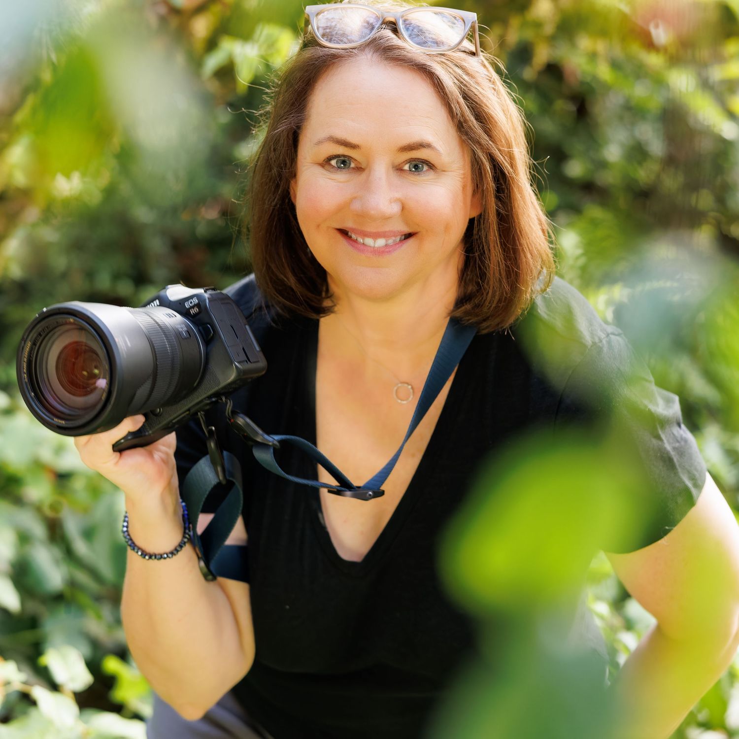 A photographer with a professional camera posing outdoors with greenery in the background.