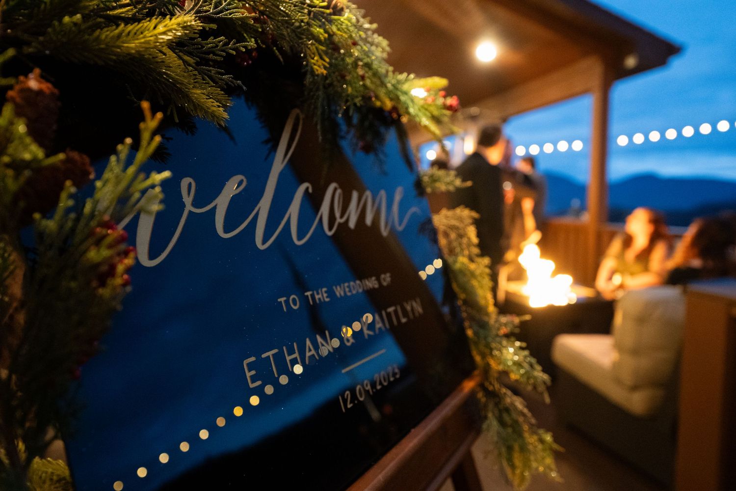 A welcome sign decorated with greenery and string lights glows warmly at an evening outdoor gathering.