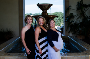 Women in formal dresses pose together in an elegant indoor setting.