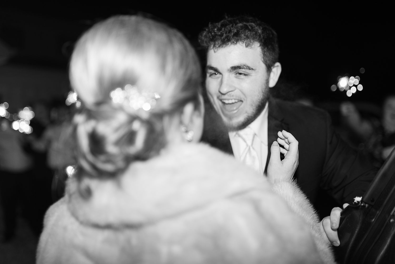 Black and white candid reception photo showing guests in conversation with festive lights.