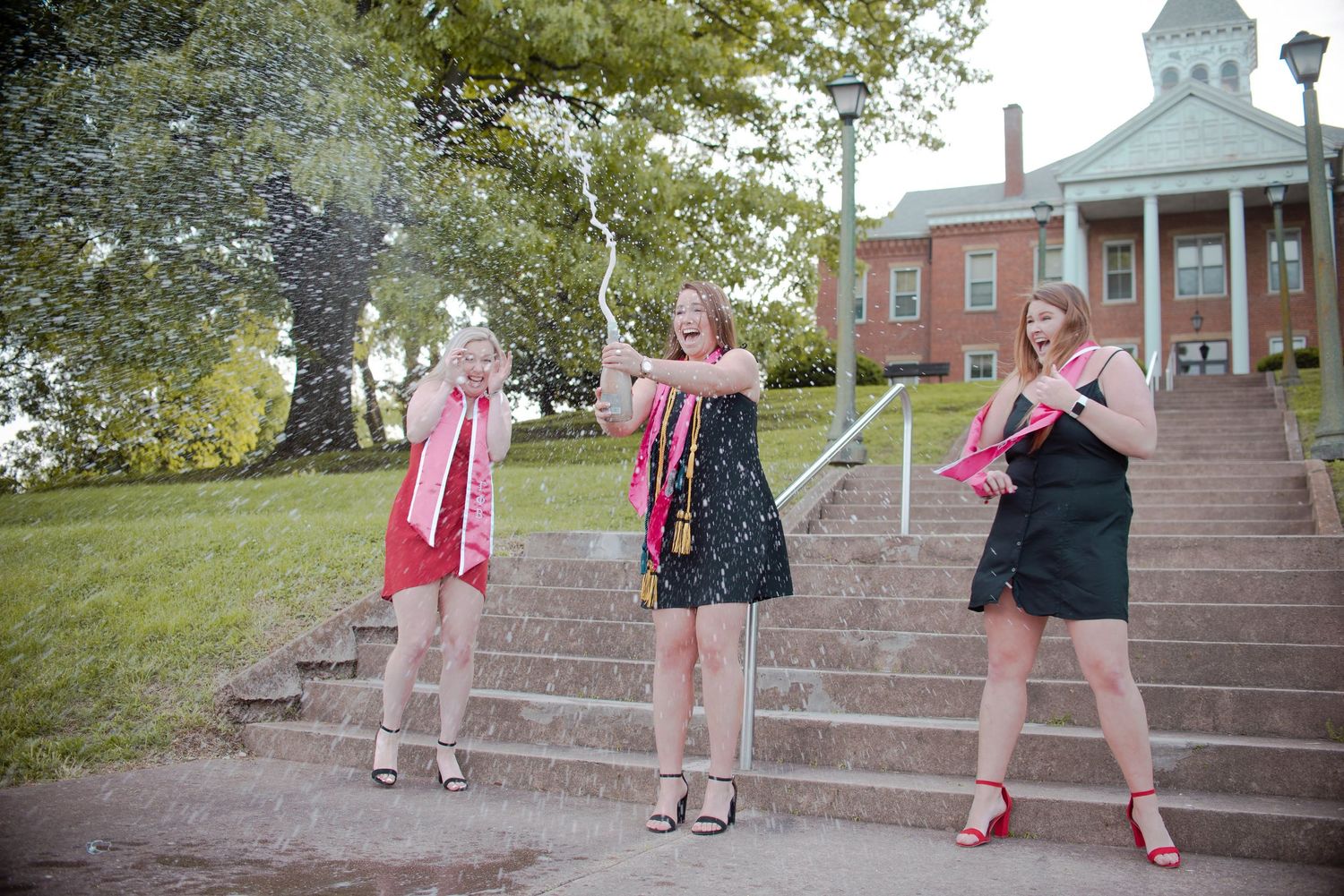 Three figures pose playfully on stone steps leading up to a red brick building with white columns.