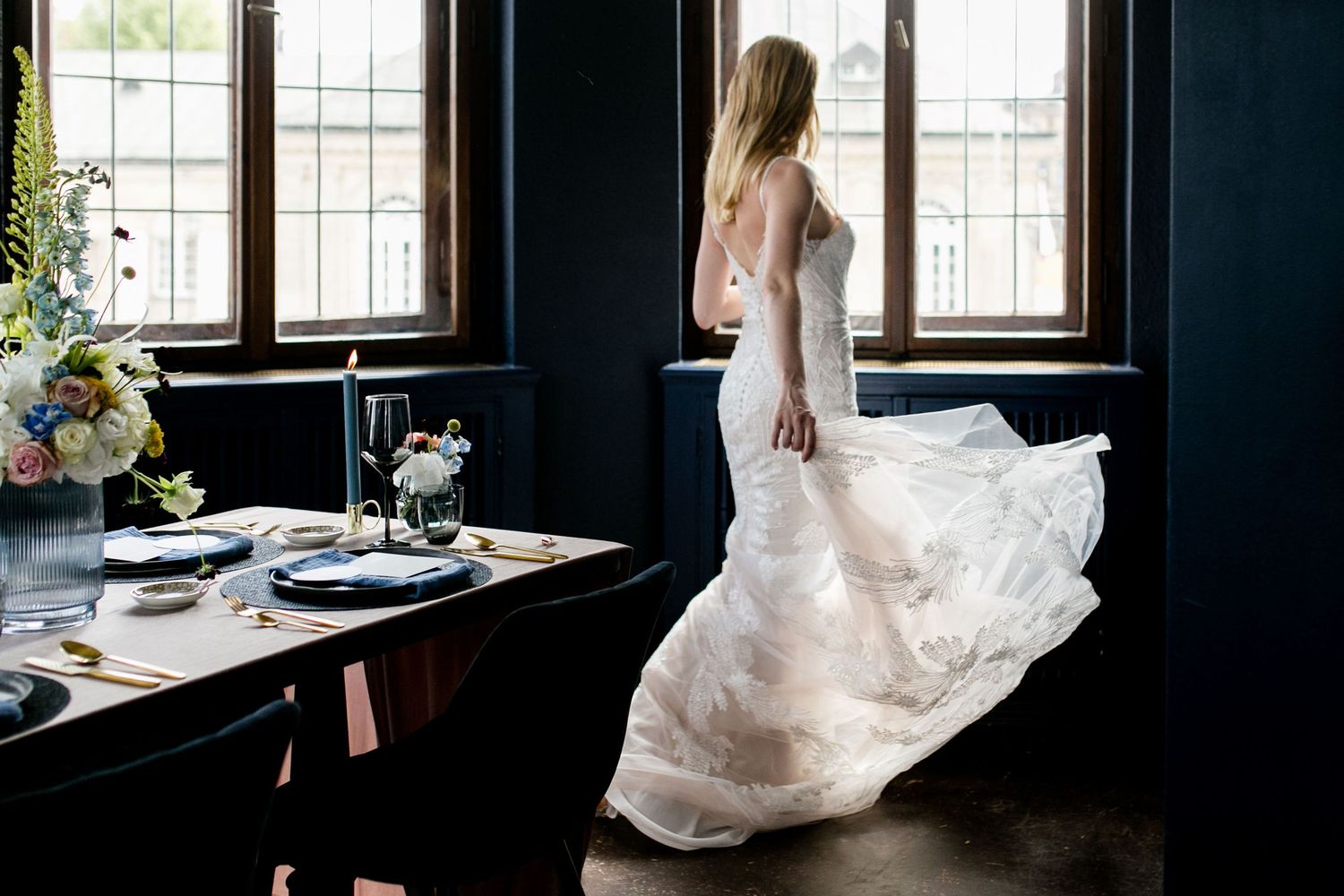 A flowing white wedding dress twirls elegantly in a naturally lit room with dark walls and large windows.