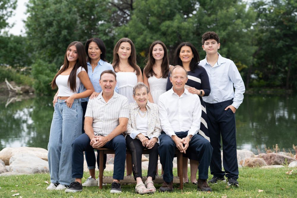 Multi-generational family portrait outdoors by a pond with trees in the background.
