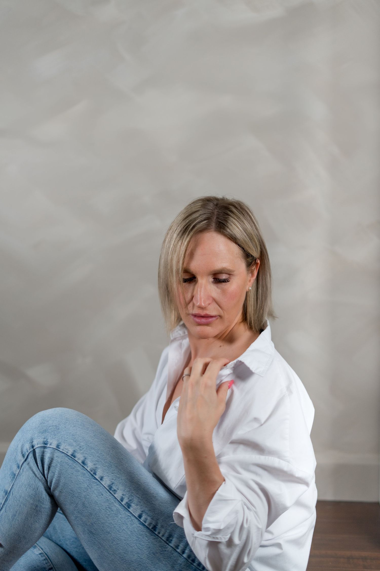 Person in white button-down shirt and jeans sitting against textured wall.