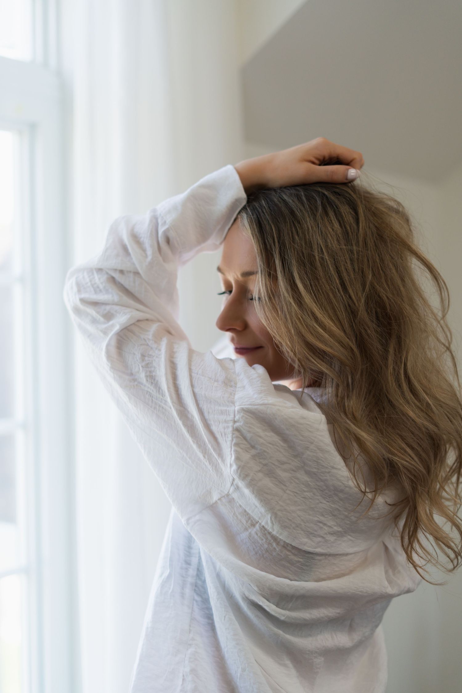Woman in white blouse gazing contemplatively by window in soft natural light.