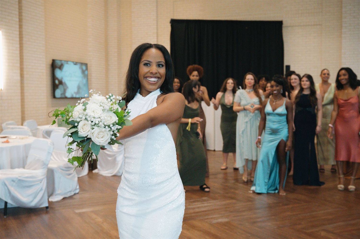 A joyful bride holds her white bouquet while guests in colorful formal dresses look on at a wedding reception.