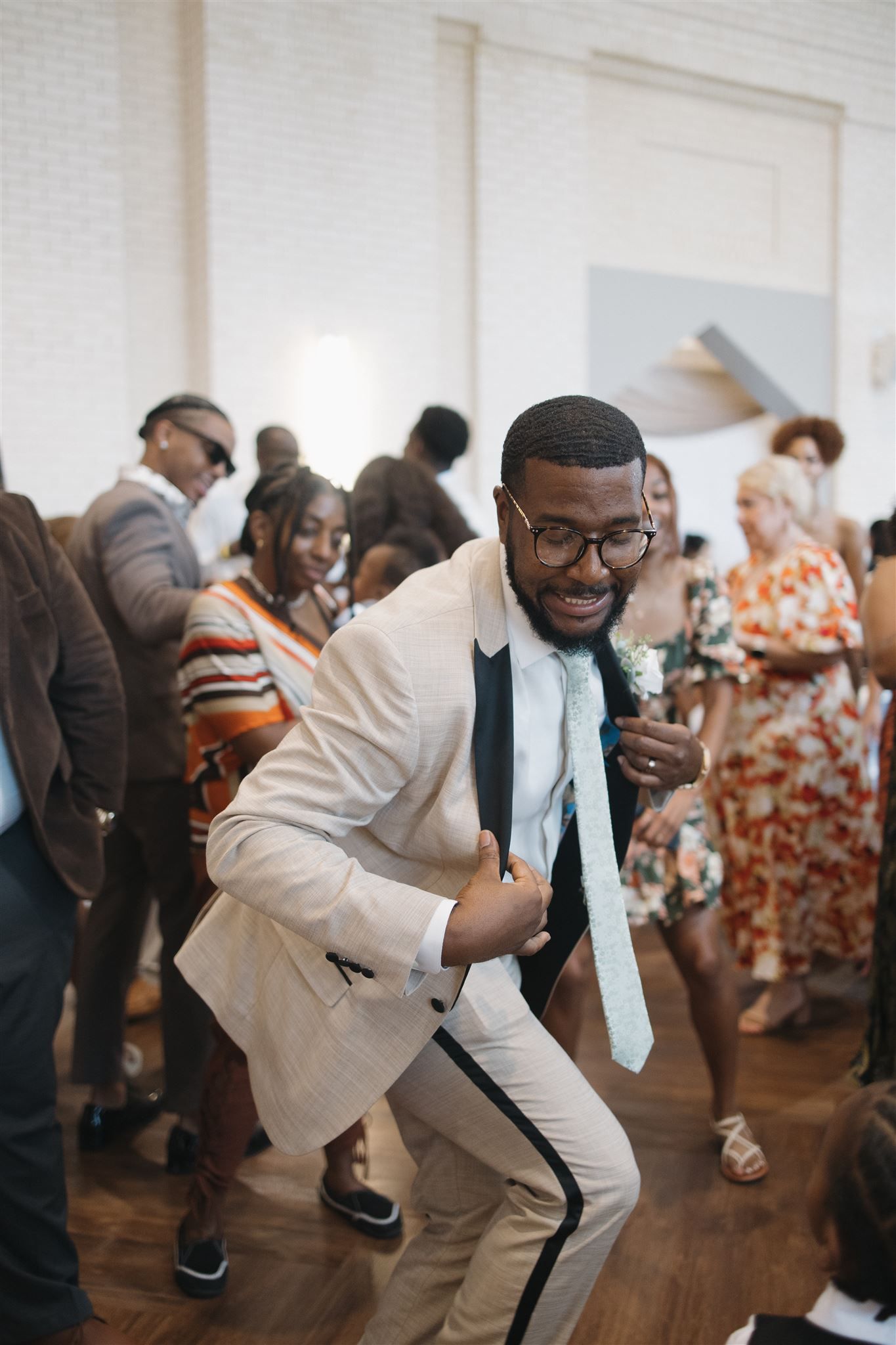 Partygoer wearing a cream colored suit and glasses dances enthusiastically at a lively indoor celebration.