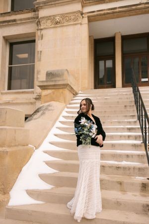 Elegant evening look featuring a white lace skirt and black shrug beside historic stone steps of a building.
