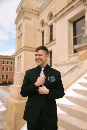 A person in a formal black suit and blue boutonniere stands smiling on stone steps outside a classic building.