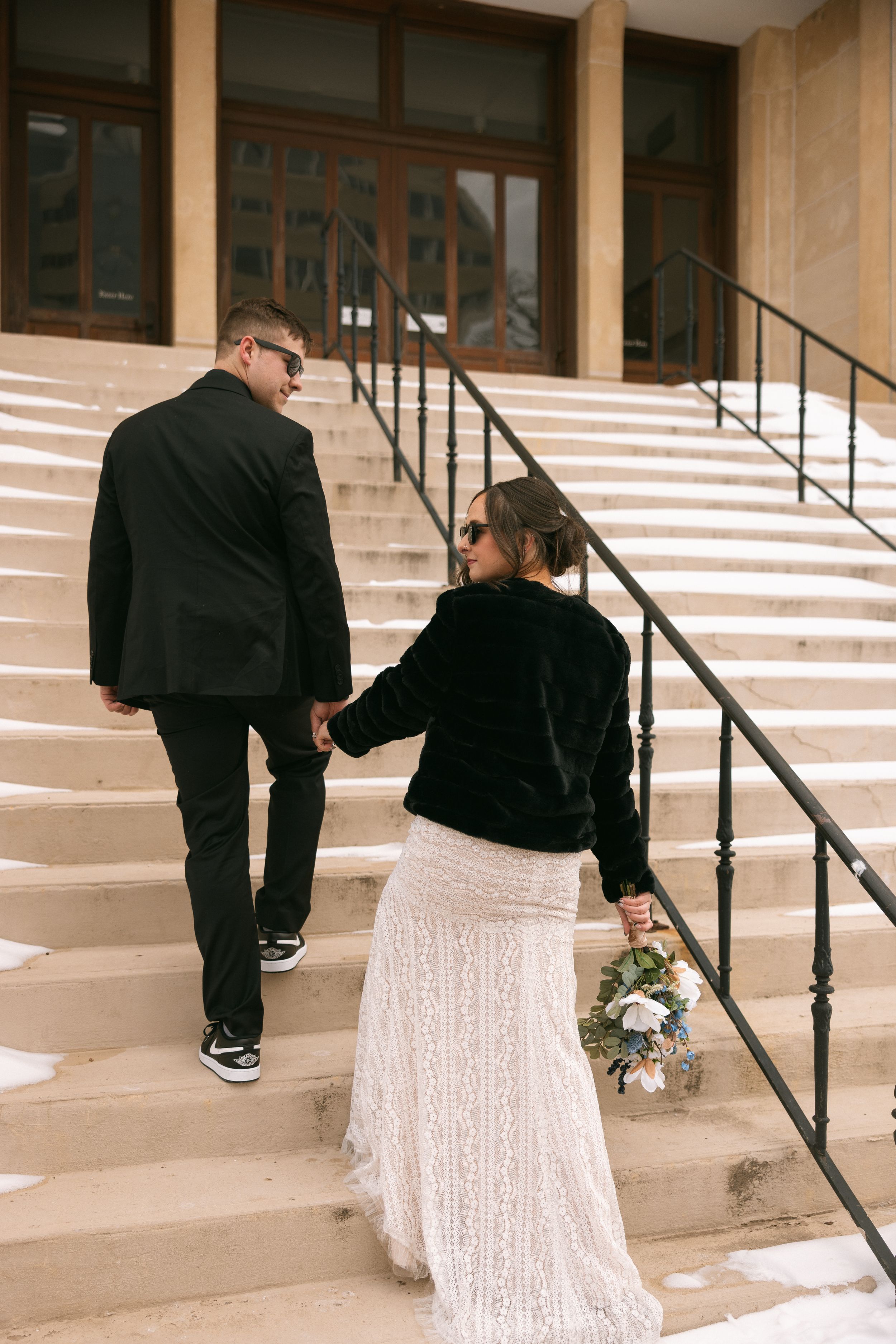 A couple on concrete steps outside a building, with the person holding a bouquet wearing a long white lace skirt.
