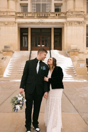 A couple in wedding attire pose on grand stone steps outside an elegant building with classic architecture.