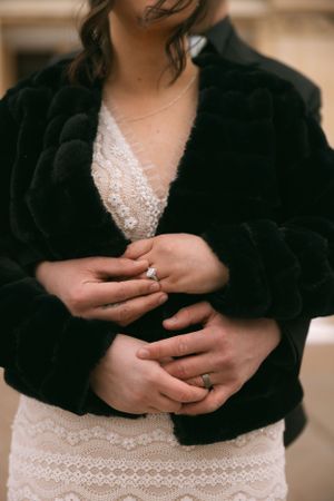 Close-up of hands with engagement rings against a white lace dress and black velvet cardigan.