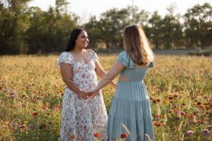 Two friends in floral and blue dresses walking together through a sunlit field of flowers.