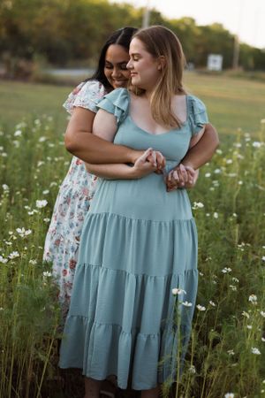 Friends in floral and blue dresses embrace in a wildflower field at sunset.