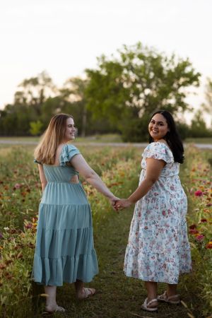 Two women in summer dresses hold hands while walking through a blooming wildflower field at sunset.