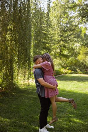 Joyful lift and embrace under weeping willow trees in a sunny garden setting.