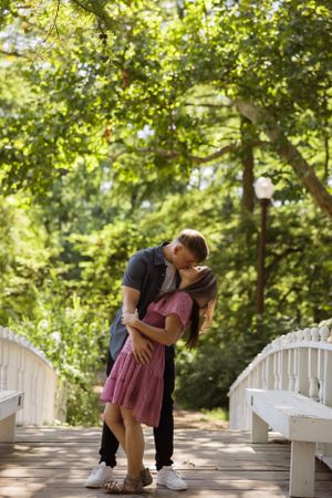 Romantic pose on white bridge with dappled sunlight filtering through trees.