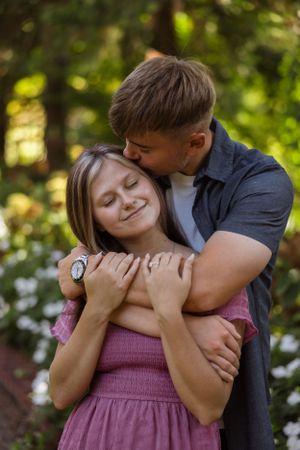 Tender embrace of couple in pink and dark clothing against lush garden background.