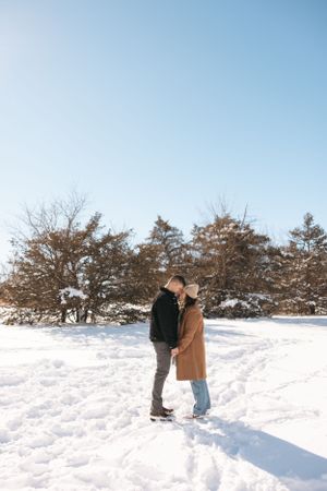 Wide shot of couple standing together in snowy field with evergreen trees in background.