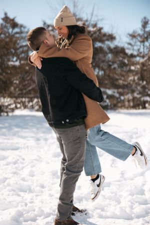 Playful winter moment as couple hugs and lifts in the snow wearing casual outerwear.