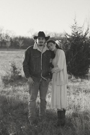 A romantic black and white photo of a couple in western attire standing together in a rustic outdoor field setting.