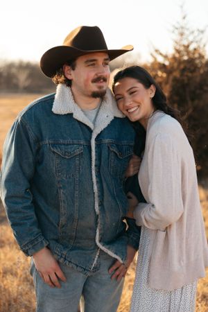 Couple embraces in a rustic setting at sunset with a cowboy hat, denim jacket, and flowy white dress.