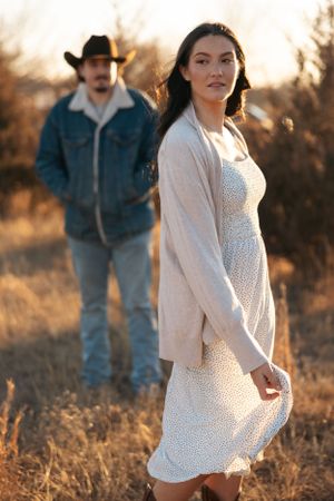 A couple poses in a field at sunset, with her in a white dress and cardigan and him wearing a cowboy hat and jacket.