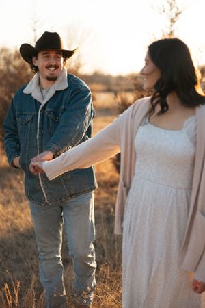 A couple holds hands at sunset in a field, with the person on the left wearing a cowboy hat and denim jacket.