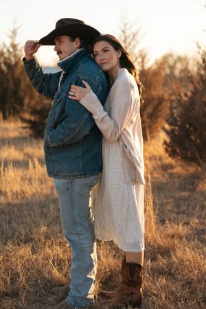 A couple stands together in a golden field at sunset, wearing country western attire and embracing.