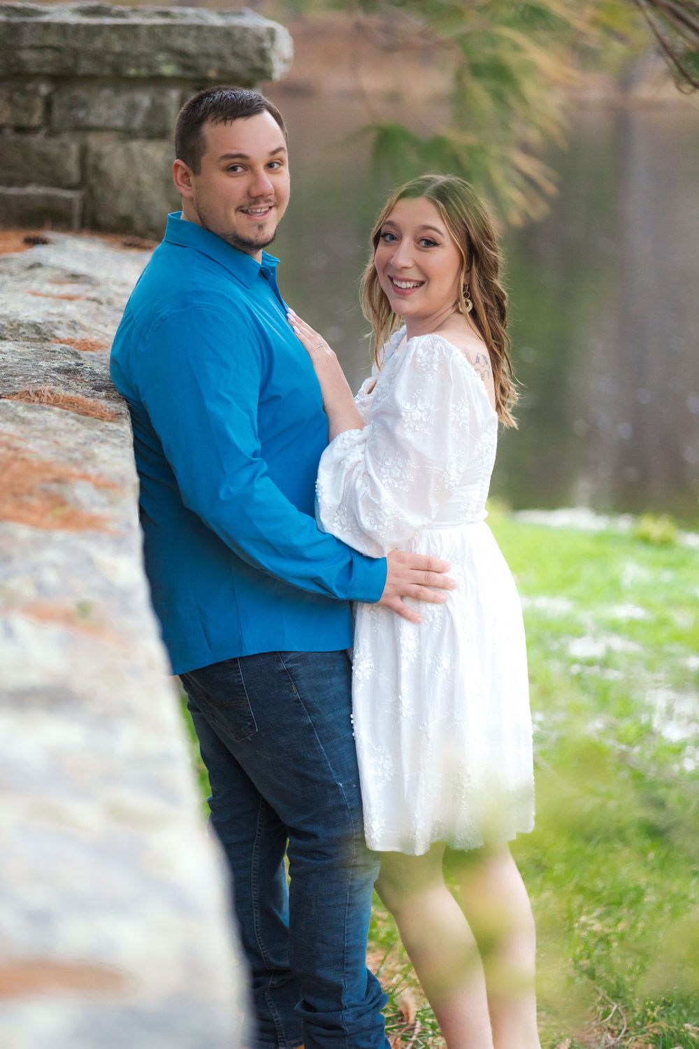 Couple poses together by stone wall near lake during outdoor photo session.