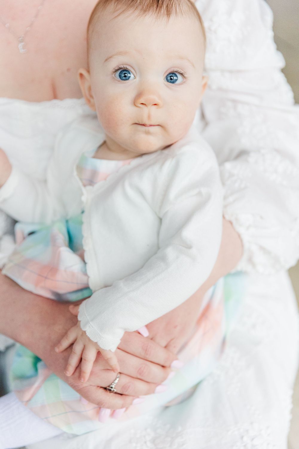 Baby in white outfit gazes at camera with bright blue eyes against light background.