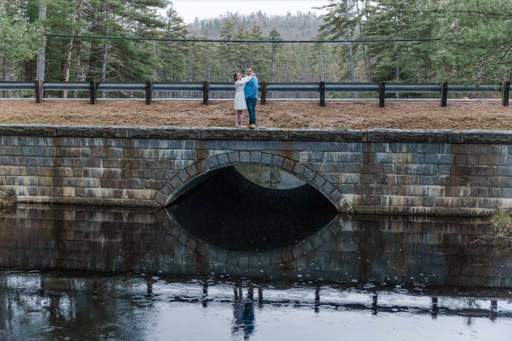 A couple stands on a stone bridge in a park surrounded by evergreen trees on a cloudy day.