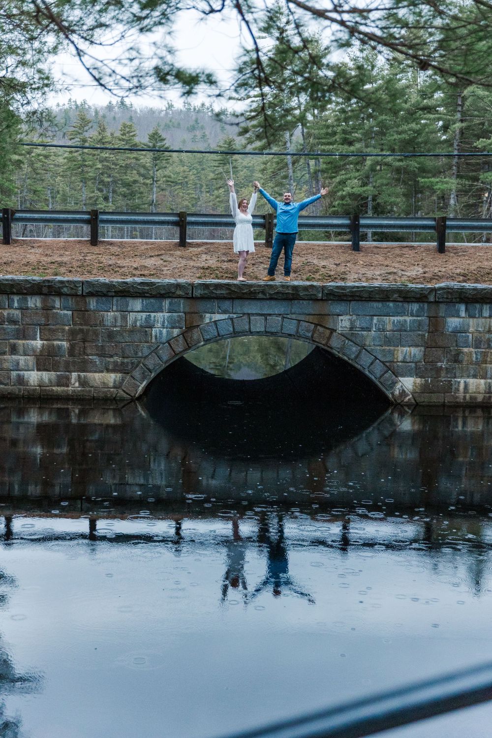 Couple stands joyfully on a stone bridge over calm water reflecting pine trees in the background.
