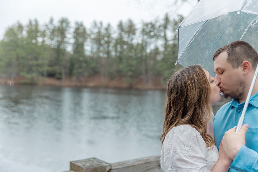 Couple shares intimate moment under clear umbrella by lakeside during engagement session.