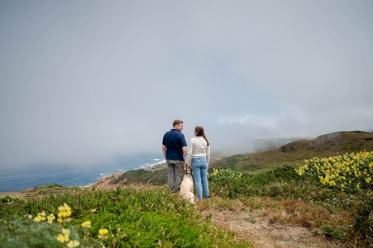 Couple standing smiling at each other with their dog along a coastal trail with yellow wildflowers and misty ocean views in the background.