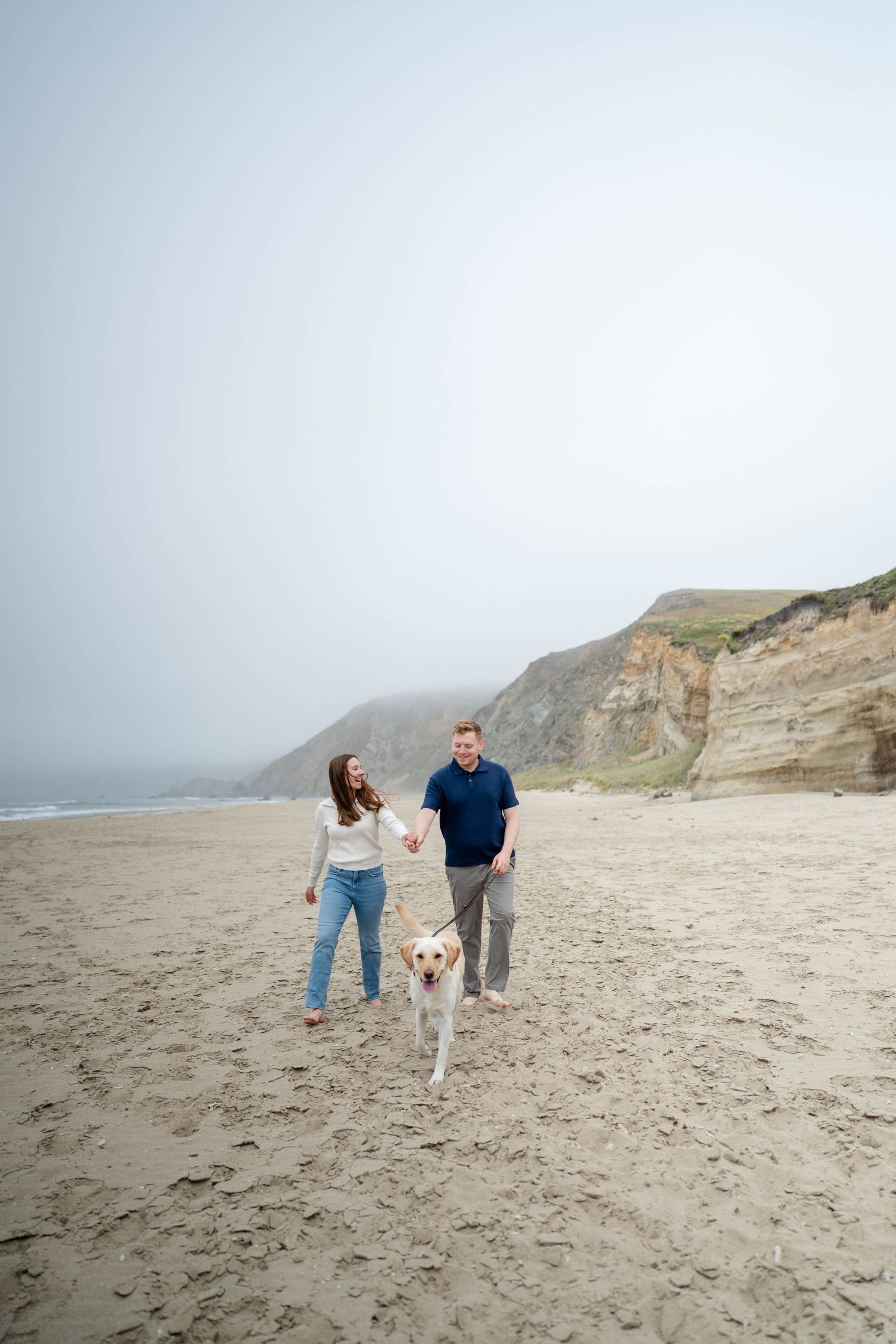 Couple walking with their golden Labrador retriever along a misty beach with cliffs in the background.