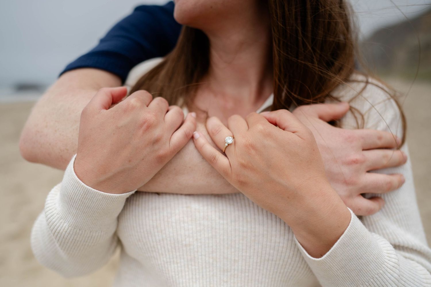 Woman wearing white sweater holds the arm of her significant other showing off her engagement ring.