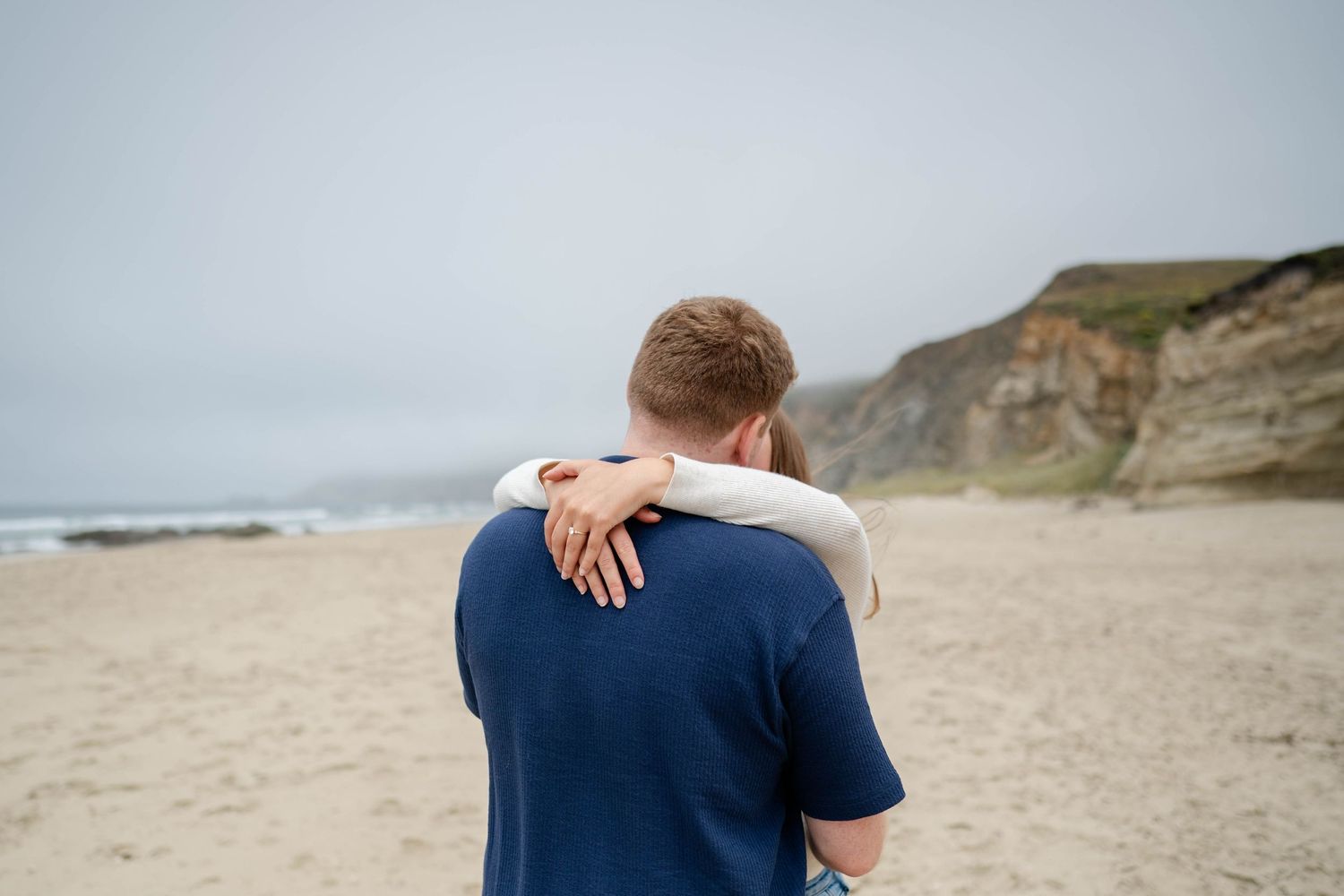 A man navy blue shirt on the beach with their back turned to the camera and their fiancé having her arms over his shoulders.