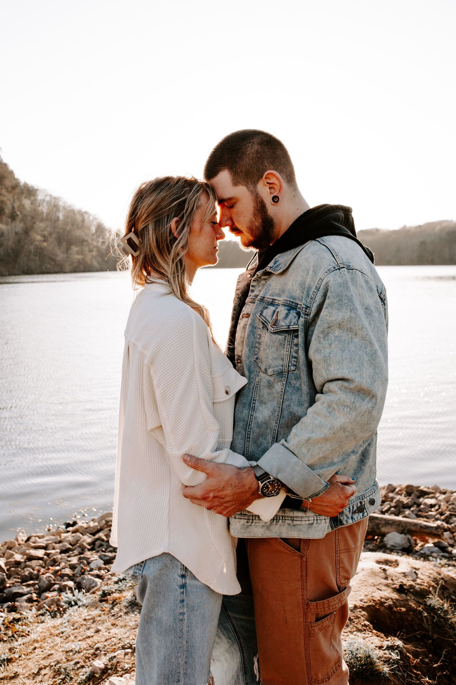 Romantic couple sharing an intimate moment by a serene lake shore on an autumn day, wearing casual denim and white clothes.