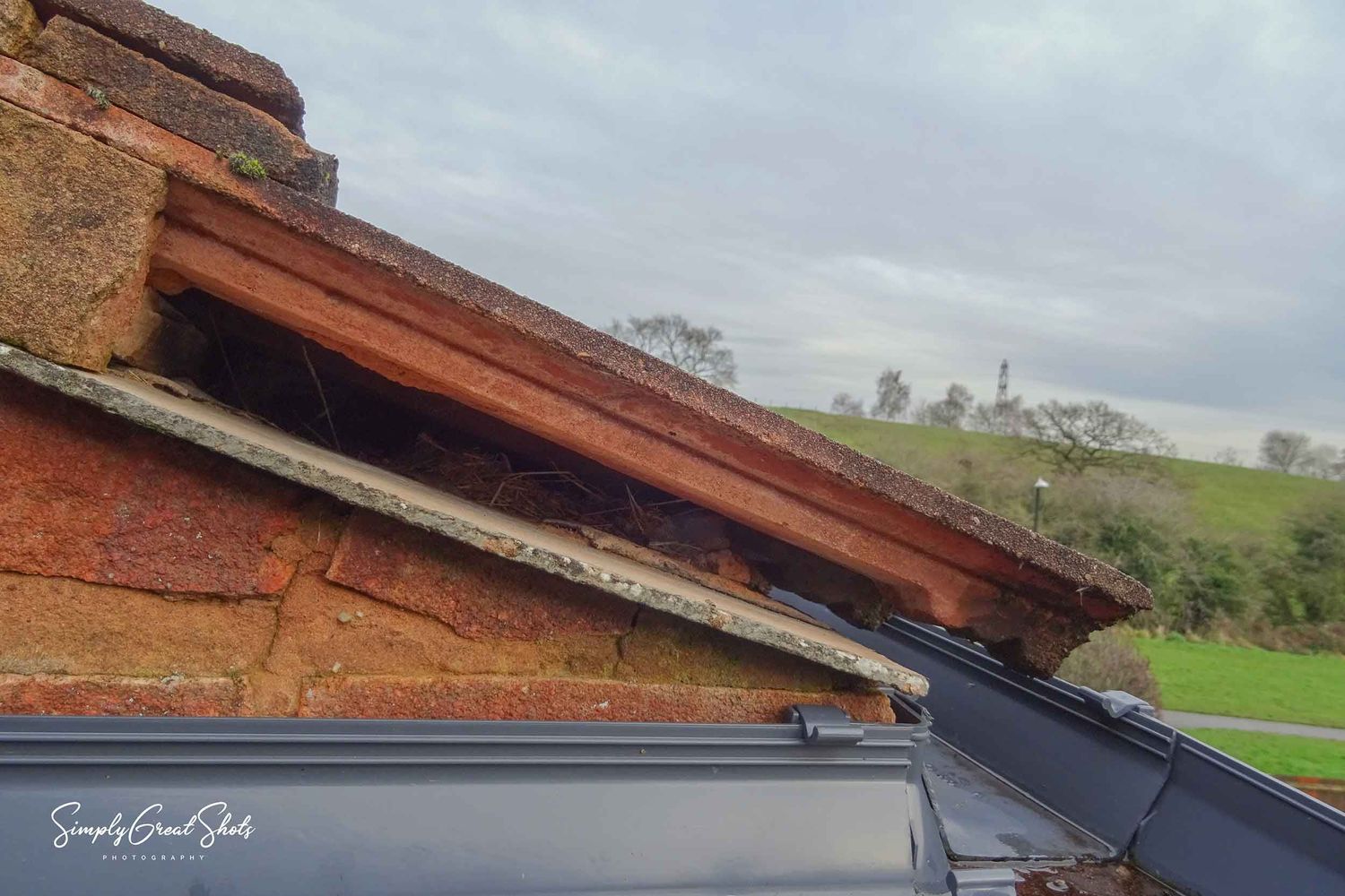 Damaged roof with rusted metal and exposed bricks against cloudy sky and green countryside in background.