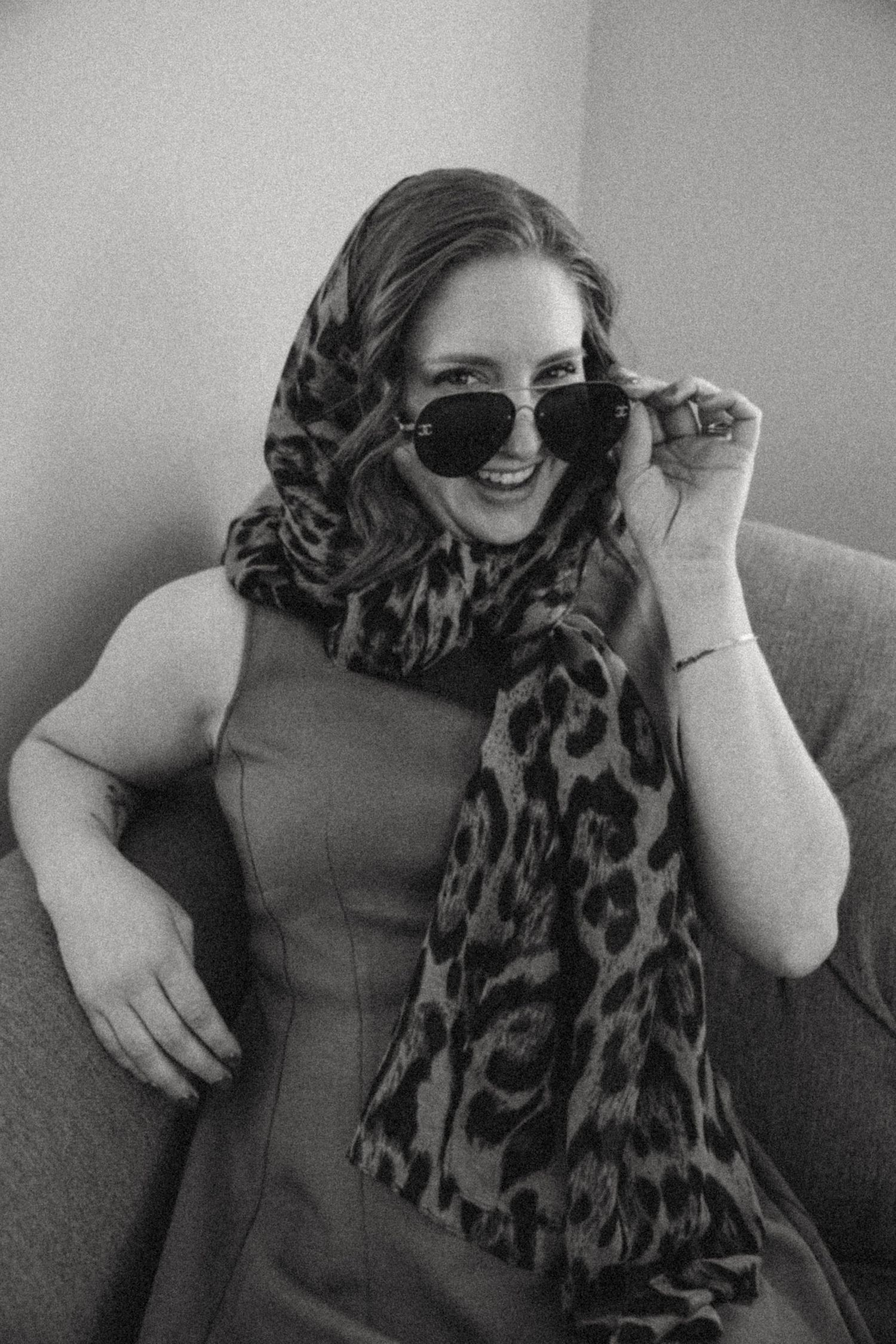 Stylish woman wearing leopard print scarf and sunglasses poses on grey couch.