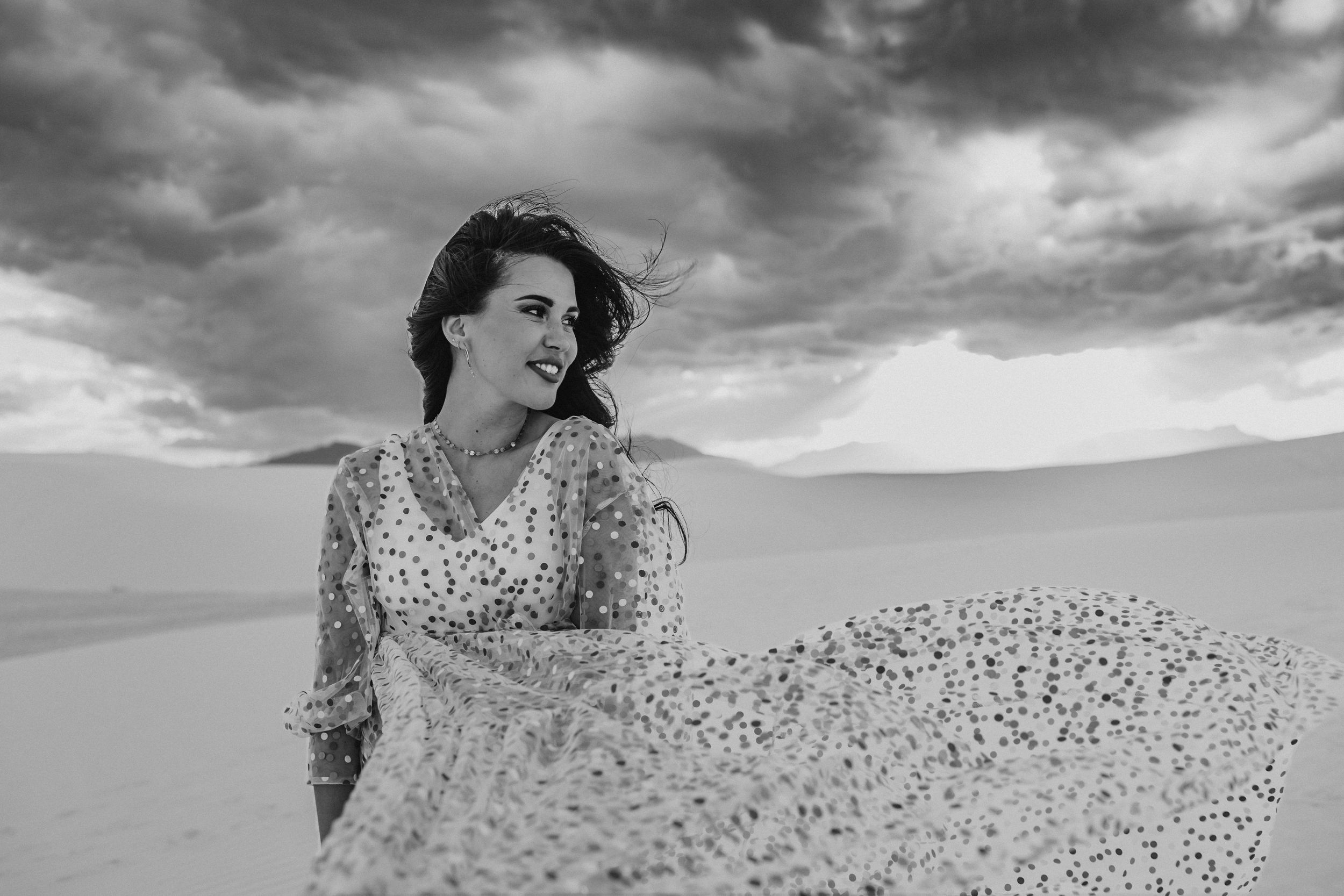 Woman in flowing dress poses dramatically against sand dunes under stormy sky.