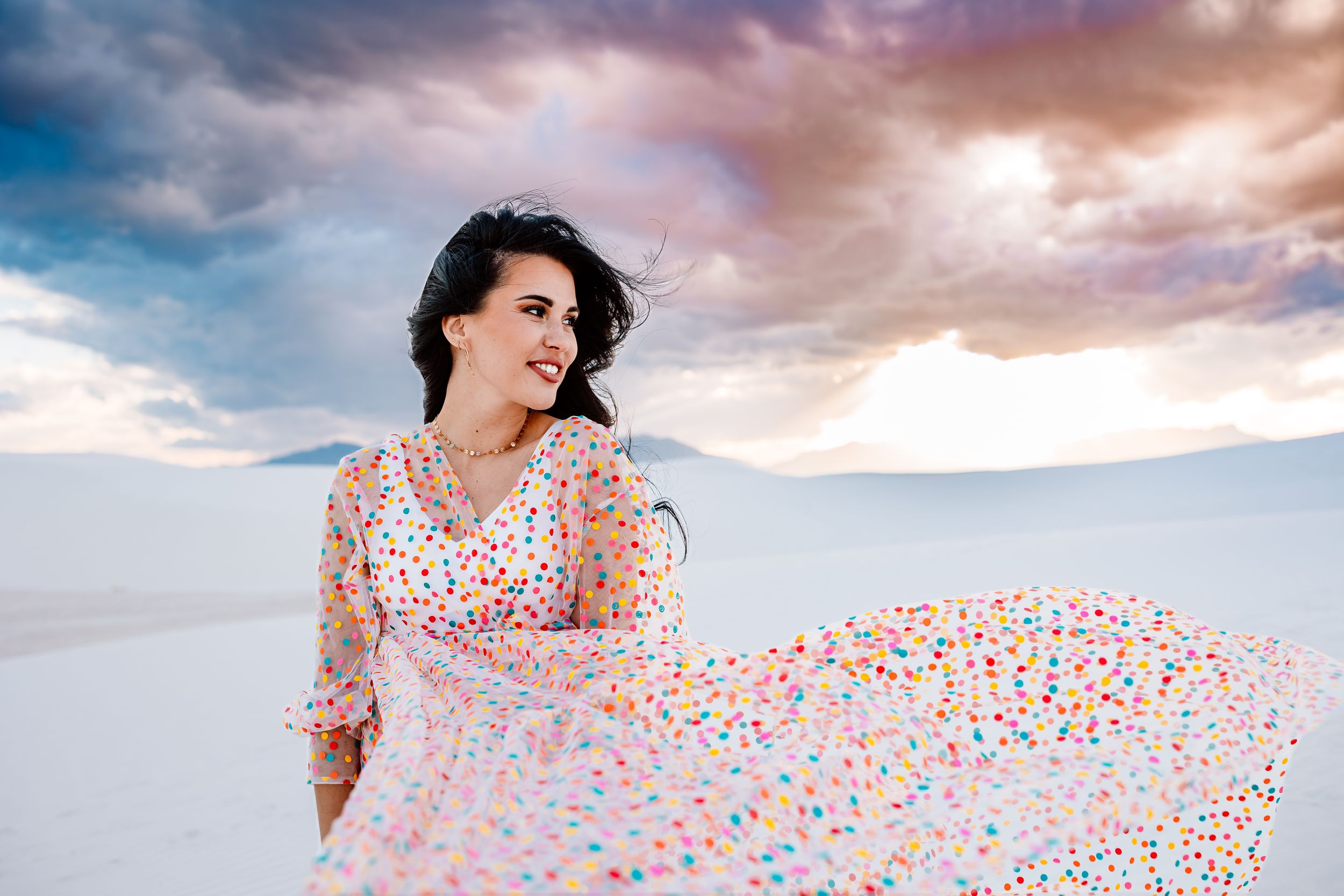 A flowy white dress with colorful polka dots twirls against a dramatic desert sky at sunset.