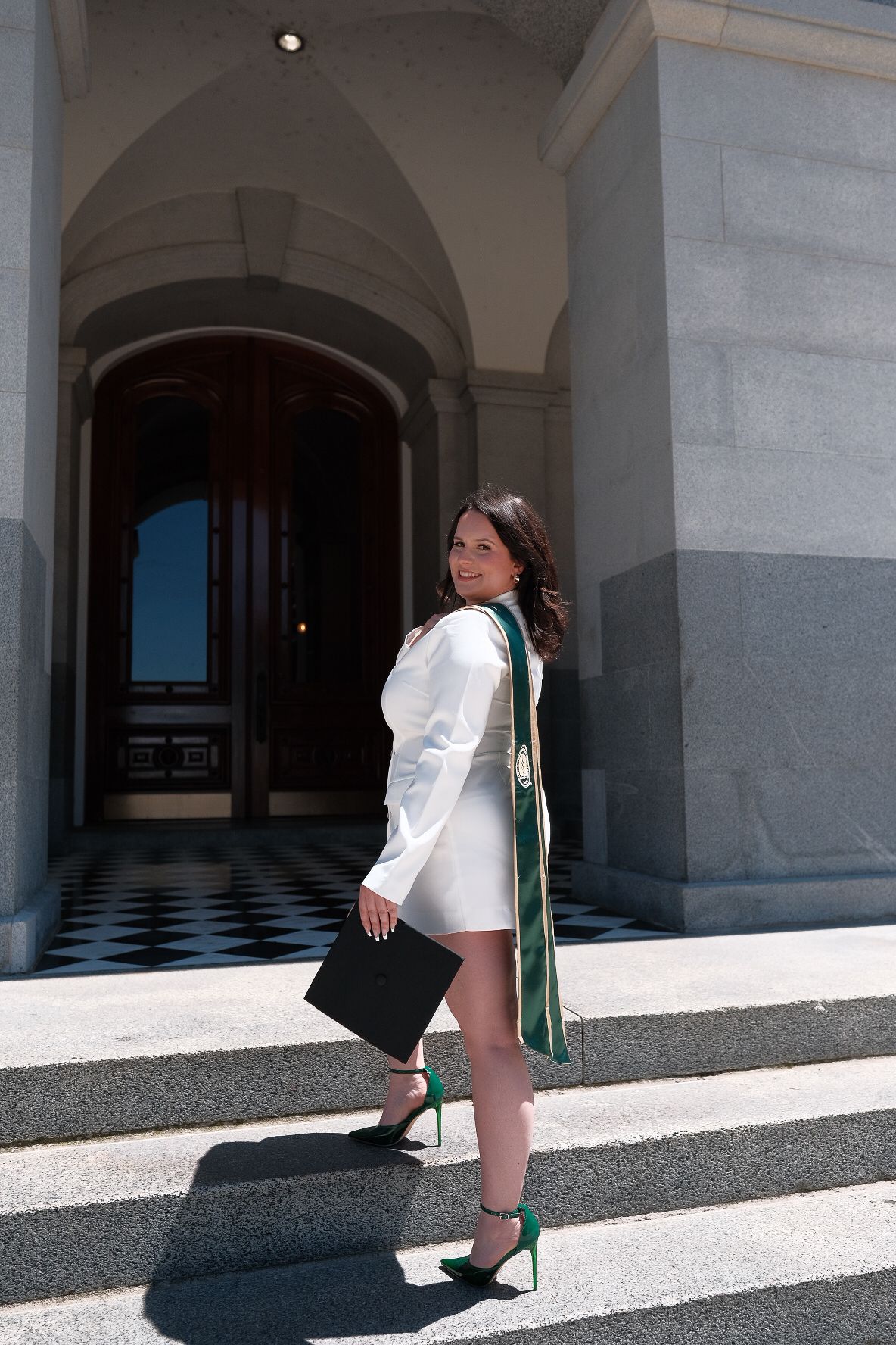 A graduate in white and green regalia poses against a checkered floor and arched doorway.
