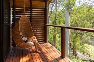 Woven egg chair hangs on wooden deck balcony overlooking lush green bush scenery in Australia