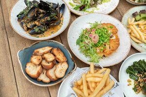 Overhead view of various restaurant dishes including mussels, french fries, shrimp, and salad arranged on wooden table.