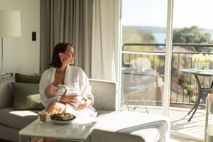 Woman in white robe relaxing on sofa with snacks, enjoying sunlit hotel room with balcony and scenic water view.
