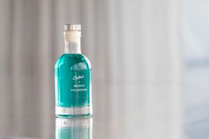 A small teal glass perfume bottle from Bronte Collection displayed on a reflective surface with soft neutral background.