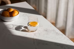 Orange in a white bowl with oranges on a sunlit table creating dramatic shadows.