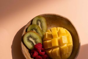 Fresh sliced mango and kiwi arranged with bright red raspberries on a brown ceramic plate against a pink background.